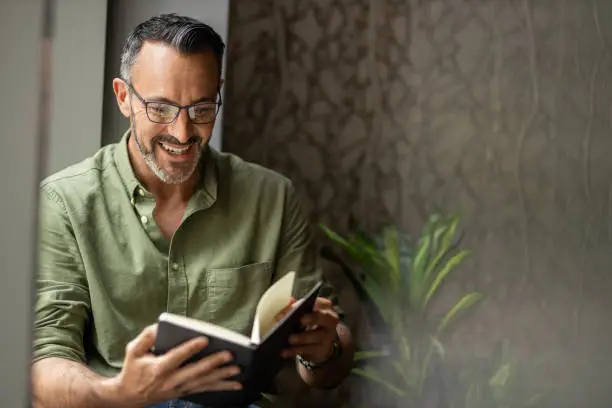 Photo of Mature man reading book by window, smiling wearing glasses, copy space