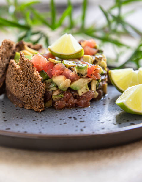 istock Raw tuna and avocado tartar served with rye bread crackers and lime on black ceramic plate. Gourmet fish tartar. 1320156779