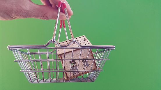 Real estate market and real estate purchase, the fingers of a person holding a house icon and a shopping cart. Buying a home concept on a green background. Copy space.
