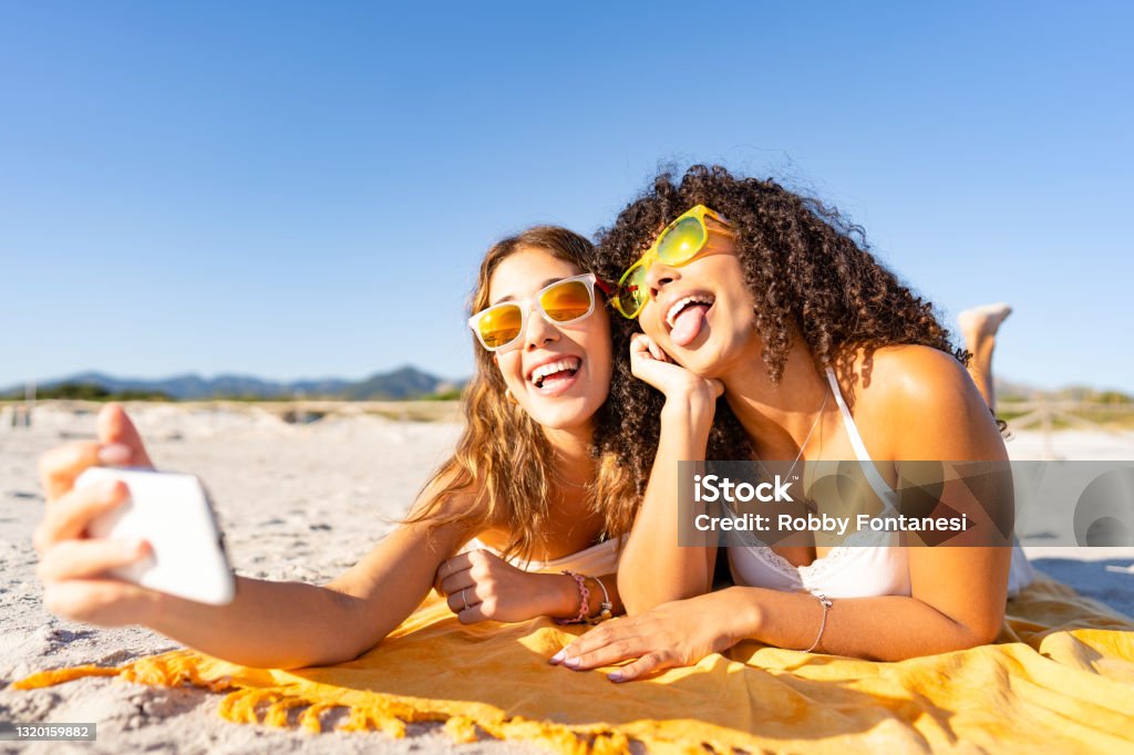 Two beautiful girls lying on the beach in the summer having fun making faces with their tongues out while taking a selfie. Two multiracial lesbian girlfriends with funny sunglasses using smartphone Beach Stock Photo