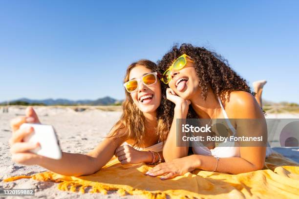 Two Beautiful Girls Lying On The Beach In The Summer Having Fun Making Faces With Their Tongues Out While Taking A Selfie Two Multiracial Lesbian Girlfriends With Funny Sunglasses Using Smartphone Stock Photo - Download Image Now