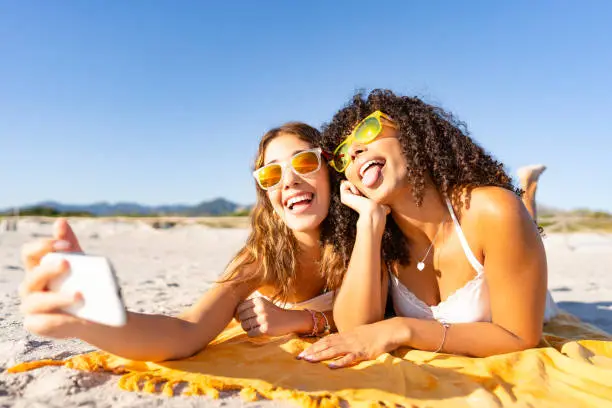 Photo of Two beautiful girls lying on the beach in the summer having fun making faces with their tongues out while taking a selfie. Two multiracial lesbian girlfriends with funny sunglasses using smartphone