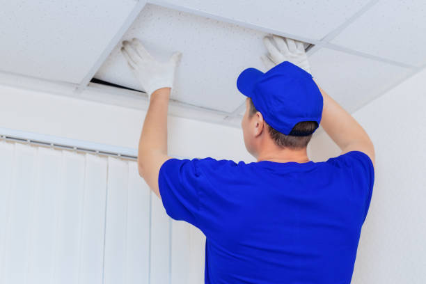 A worker in overalls installs a ceiling tile made of drywall in an office space