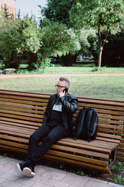 istock Young caucasian man in black jeans, leather jacket and with backpack, sits on bench in city park and talks on smartphone. Rest in an urban environment, city life. 1320168351