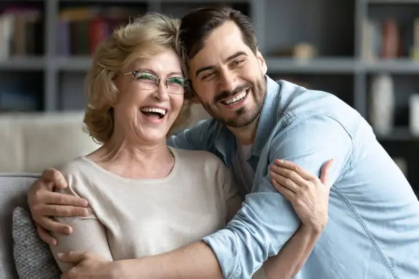 Happy middle aged retired woman relaxing on sofa with grown up handsome son, watching funny movie on tv, laughing at jokes at home. Affectionate young man cuddling older mother, having fun on weekend.