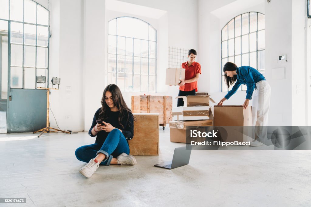 A young adult woman is looking her smartphone in a new loft office A young adult woman is looking her smartphone in a new loft office. She's taking a break during the move. New home or office concept. Two other colleagues in the background. Business Stock Photo