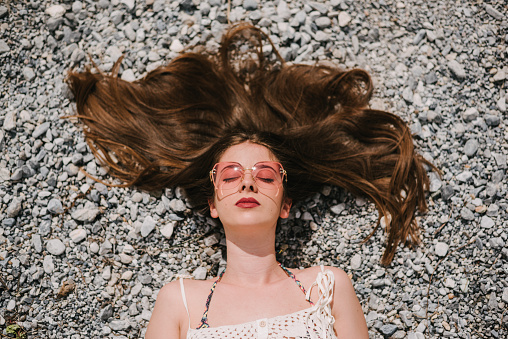 A close-up of a young adult eyes-closed woman with disheveled hair lying and relaxing on the beach with sunglasses