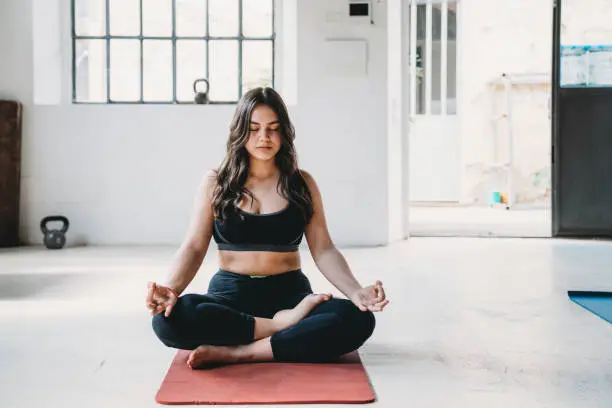 Young adult woman is practicing yoga in a modern loft. Half lotus or ardha padmasana pose.