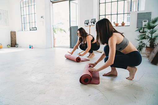 Two women are rolling their exercise mats. They are getting ready for the yoga class in a stylish loft.