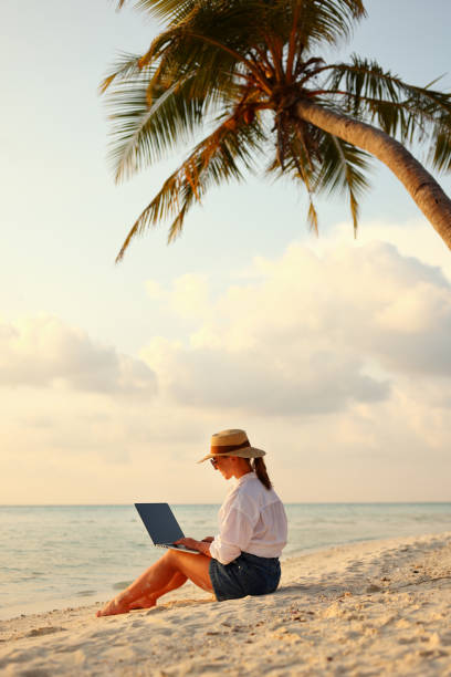 istock Young female freelancer wearing straw hat working on laptop while sitting on tropical beach at sunset 1320177906
