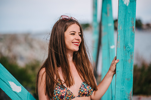 A close-up of a young adult woman standing next to a blue wooden pier holding onto it looking away smiling