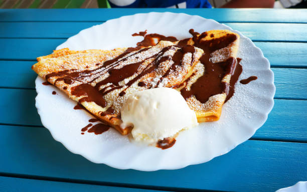 Pancake with vanilla ice cream and decorated with chocolate syrup in white plate on blue wooden table.