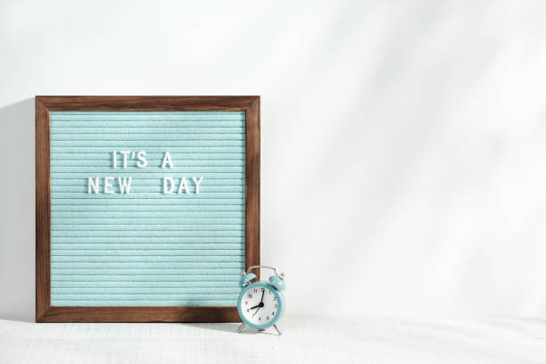 istock It's a new day. Alarm clock and letter board with a motivating phrase stand on the table. Morning sunlight with shadows. Eight o'clock in the morning. 1320186495