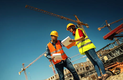 Male and female workers looking at checklist of daily jobs, talking about work schedule with construction site of a multi-story apartment building in background\n\nNOTE: blueprints are imaginary, printed solely for the purpose of shooting.