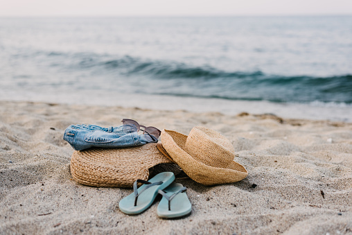 A close-up of beach accessories - straw hat, straw bag, flip-flops, denim shorts, pink sunglasses on the sand beach with the sea waves in the background