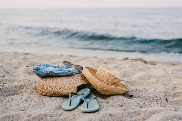 Beach accessories on the sand beach A close-up of beach accessories - straw hat, straw bag, flip-flops, denim shorts, pink sunglasses on the sand beach with the sea waves in the background straw hat photos stock pictures, royalty-free photos & images