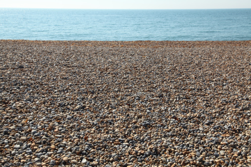 Horizontal image of a pebble beach, sparkling blue sea and pale sky for use as a background