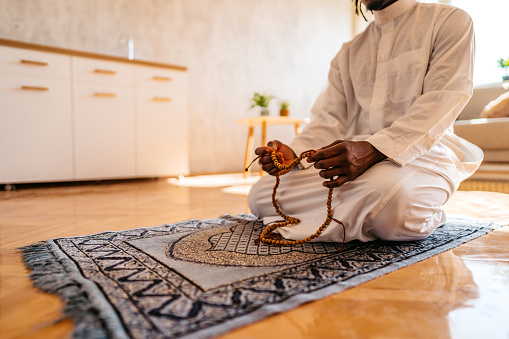 Young African-American Muslim man wearing kaftan holds prayer beads while praying at home.