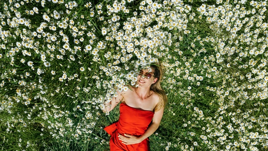 Young beautiful woman lying down in daisies meadow on a sunny day. Smiling, wearing red dress and sunglasses. Enjoying sunlight and fresh air and connecting with nature.