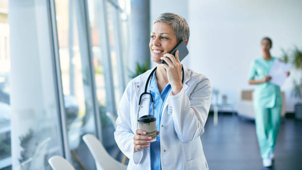 Happy healthcare worker communicating over mobile phone during her coffee break at medical clinic. Happy female doctor drinking coffee and talking on cell phone on a break in hospital. doctor utensils stock pictures, royalty-free photos & images