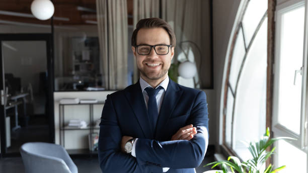 Head shot smiling businessman wearing glasses standing in office Head shot smiling confident young businessman executive wearing glasses and suit standing in modern office room, happy successful entrepreneur employee intern with arms crossed looking at camera bossy photos stock pictures, royalty-free photos & images