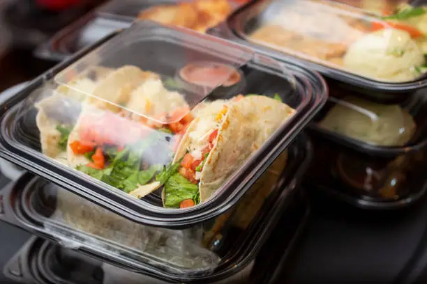 A view of several entrees prepared inside to-go plastic containers, ready for take out orders, in a restaurant setting.