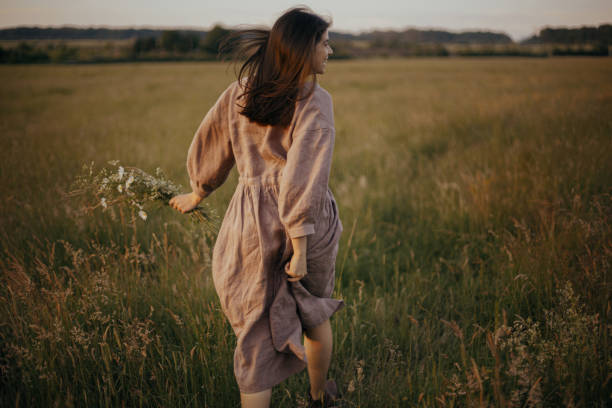 Beautiful woman in linen dress running with wildflowers in hand in summer meadow in sunset. Carefree stock photo