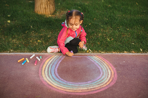 A young Caucasian girl sits on the path outside of her house chalking rainbows. She is wearing multi colored clothes with a matching tutu and has pretty jewels decorating her forehead.