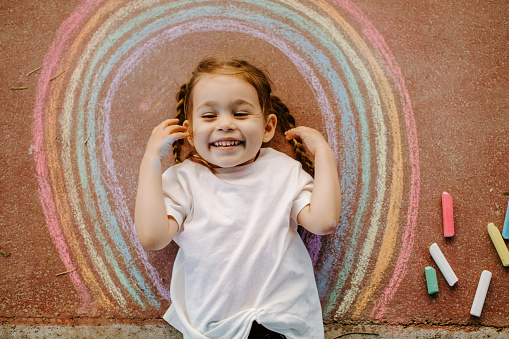 Happy little toddler girl in rainbow painted with colorful chalks on ground or asphalt in summer. Cute child having fun. creative leisure