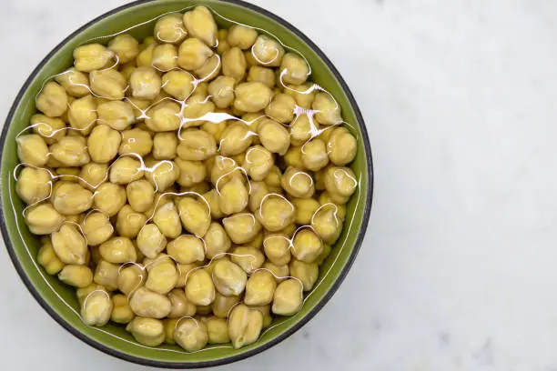 Photo of Chickpeas soaked in water, in bowl, on marble table. Concept of healthy eating, veganism, vegetarianism, meat and egg replacement. Prepare chickpeas before making hummus and other legume dishes