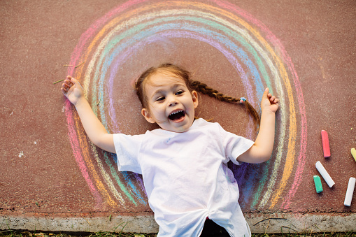 Happy little toddler girl in rainbow painted with colorful chalks on ground or asphalt in summer. Cute child having fun. creative leisure