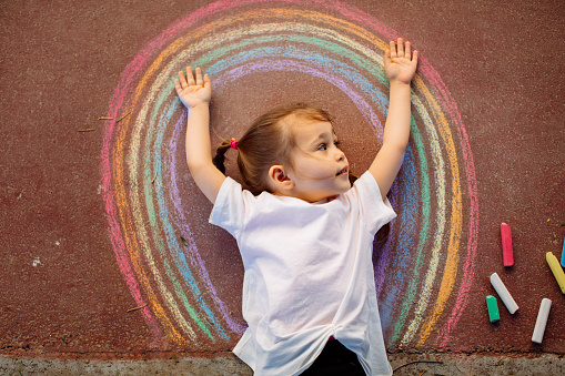 Happy little toddler girl in rainbow painted with colorful chalks on ground or asphalt in summer. Cute child having fun. creative leisure