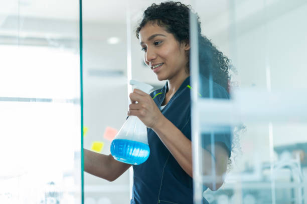 Afro-Latino woman cleans the windows of an office she works for Afro curly-haired Latin woman dressed in a blue work uniform, approximately 25 years old, an office cleaner with the help of a spray bottle, cleans the office glasses in a highly concentrated manner afro latinx ethnicity stock pictures, royalty-free photos & images