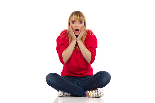 Shocked young woman in red sweater sits on a floor and holds head in hands. Front view. Full length studio shot isolated on white.