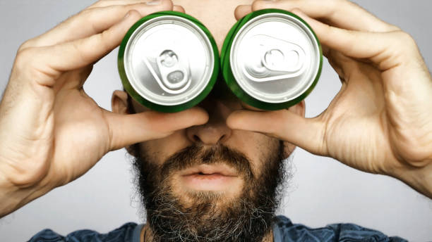 수염을 기리는 한 젊은 남자가 쌍안경을 들여다보는 척하는 맥주 두 캔을 눈에 안고 있습니다. - 30s male humor close up 뉴스 사진 이미지