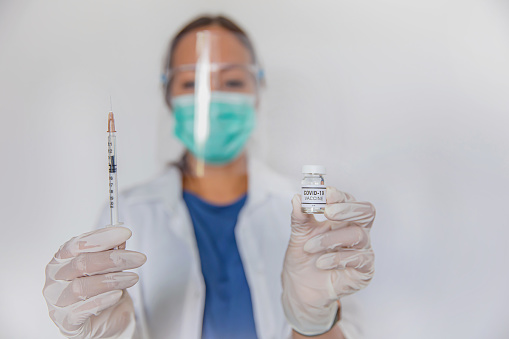 Close-up portrait shot of Asian female doctor holding syringe and COVID-19 vaccine in her hands. The doctor wearing a face shield, surgical mask and surgical gloves for safety protection.