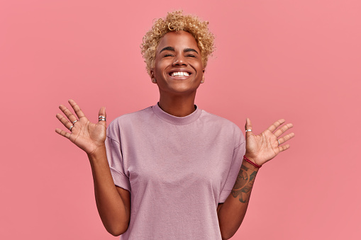 Optimistic black woman raises palms and close eyes from joy, happy to receive awesome present from someone or rejoicing triumph feeling winner, smiles broadly, in a casual lilac t shirt, on pink wall.