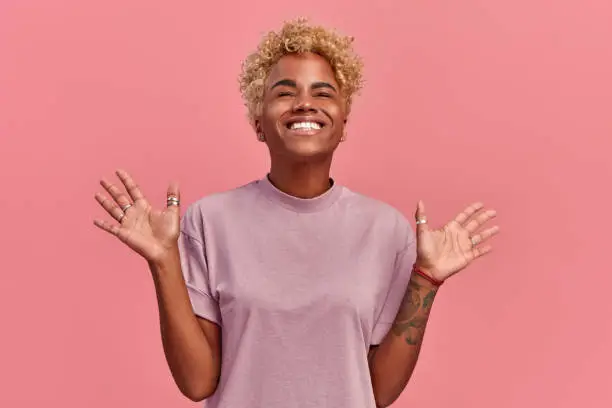 Photo of Optimistic black woman raises palms and close eyes from joy, happy to receive awesome present from someone or rejoicing triumph feeling winner, smiles broadly, in a casual lilac t shirt, on pink wall