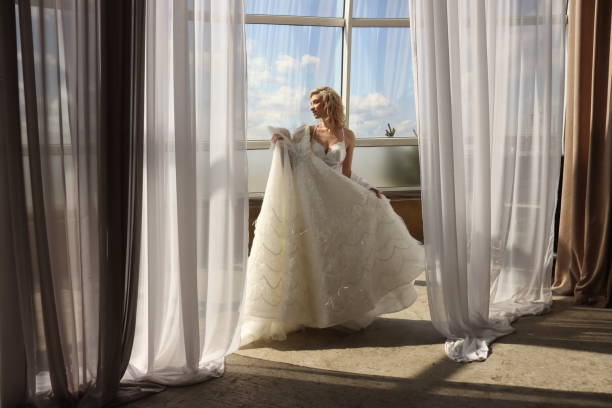 istock Bride is preparing for wedding, examines her wedding dress on background of large panoramic window. Blonde girl with white dress of bride. Blonde girl with white dress of bride 1320265906