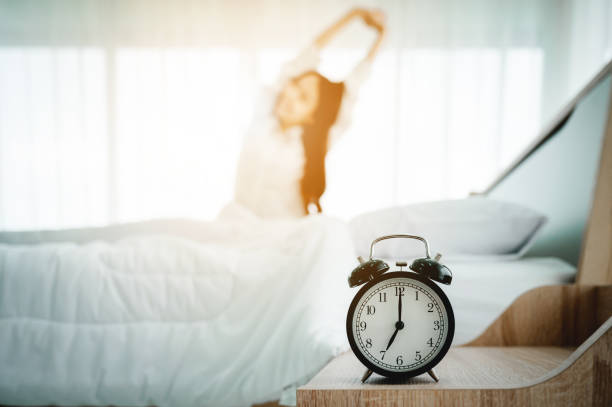 おはようございます新しい日。目覚まし時計が目を覚まし、寝室の窓の横のベッドに座っている女性の体
