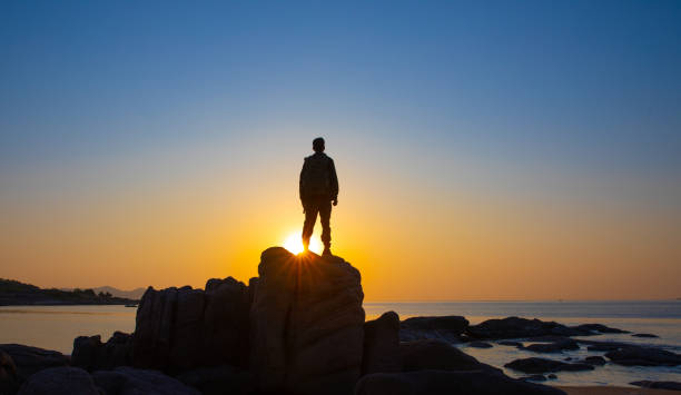 istock Young man standing on rock looking at sea sunrise 1320275625