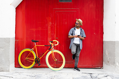 Hispanic man against an old red door holding his phone beside his vintage bicycle. Sustainable tourism and mobility concept