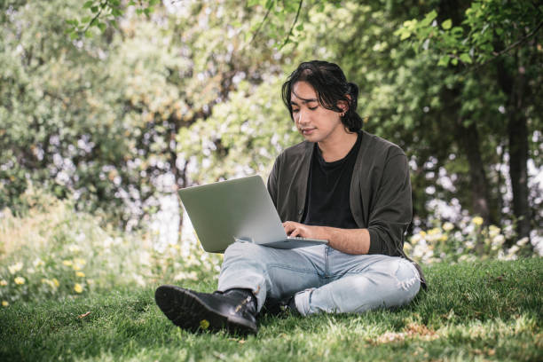 istock Young man works remotely using his laptop sitting on the grass at a park in springtime. 1320291360