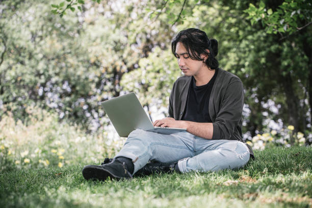 istock Young man works remotely using his laptop sitting on the grass at a park in springtime. 1320291580
