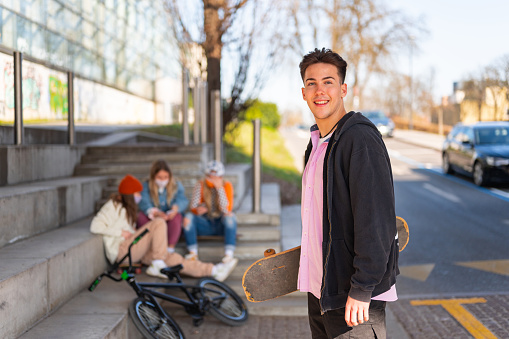 Portrait of a young man holding a skateboard outdoors. Small group of people sitting in the background.