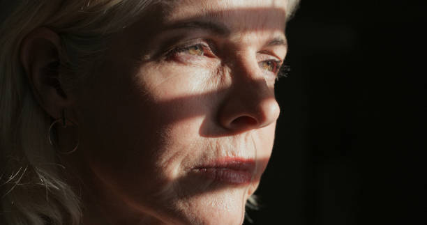 Shot of a mature woman sitting alone in a dark room at home and looking outside contemplatively This is not how I wanted things to go human face light stock pictures, royalty-free photos & images