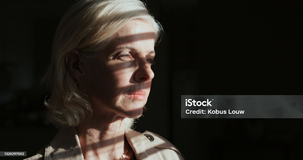 Shot of a mature woman standing alone in a dark room at home and looking outside contemplatively This is the best room for me to think in Guilt Stock Photo