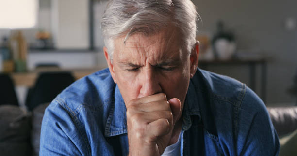 Shot of a mature man sitting alone on the sofa at home and coughing I think I'm getting a chest infection coughing photos stock pictures, royalty-free photos & images