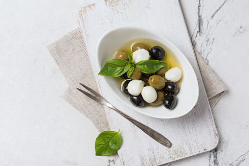 Top view of tasty mix green and black olives with mozzarella, basil leaves on white wooden board and marble background