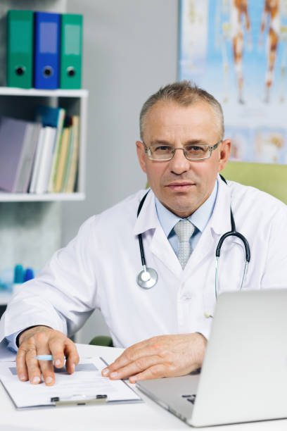 istock General practitioner looking at camera, posing in office. Confident old mature male head doctor physician in white medical uniform in glasses sitting at workplace 1320309703